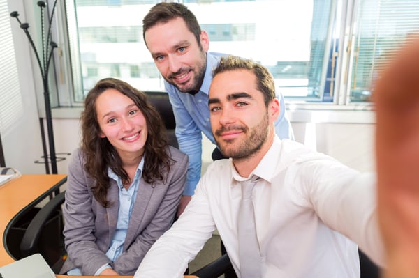 Ein Gruppenfotos als Selfie in einem Büroraum, zeigt mehrere Personen, die lächelnd in die Kamera schauen, umgeben von Büromöbeln und Arbeitsplätzen