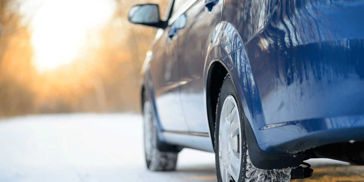 Blaues Auto im Winter, von links aus niedrigem Blickwinkel fotografiert, umgeben von Schnee, mit Sonnenschein im Hintergrund, der Bäume und den klaren Himmel beleuchtet.