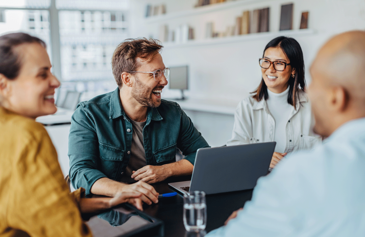 Ein Team von Mitarbeitern in einem hellen, freundlichen Büro, die sich über ein Projekt austauschen. Ein Laptop auf dem Bürotisch, um sich zum Thema Vorsorge und Arbeitgeber-Attraktivität und eine positive Unternehmenskultur austauschen.